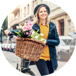 A young woman with blonde hair, wearing a black hat, a yellow sweater, and a blue coat, is smiling while walking her bicycle down a city street. The bicycle has a wicker basket filled with pink flowers.