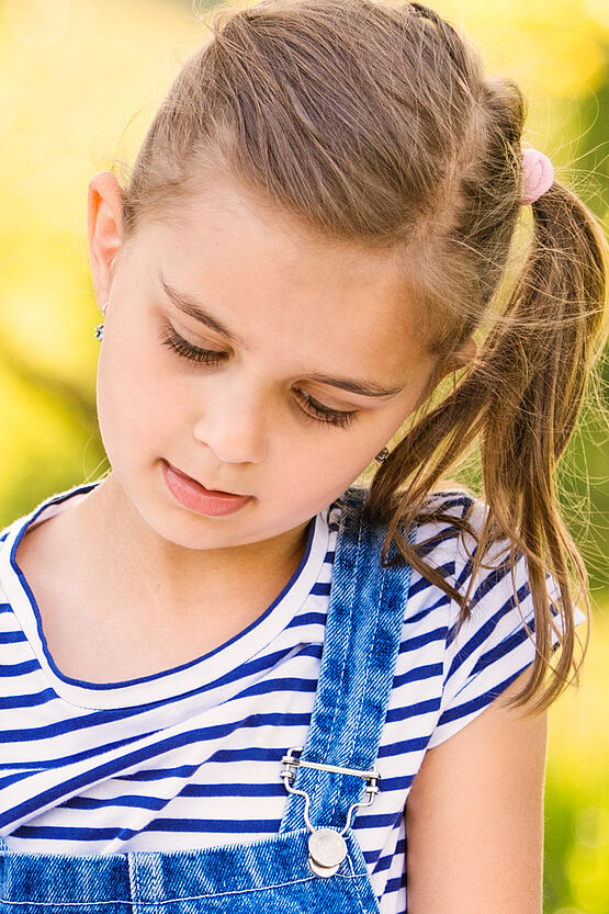 Young girl with a ponytail looking down thoughtfully, wearing a striped shirt and denim overalls, in a sunny outdoor setting